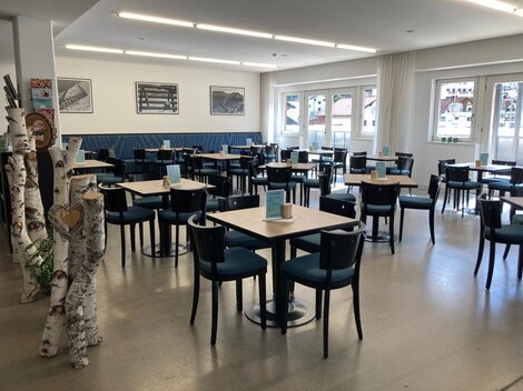 A modern restaurant with tables and chairs. The walls are bright and large windows let in plenty of light.