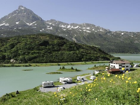 A picturesque landscape with a calm lake and an impressive mountain in the background. At the shore, RVs and a guesthouse are surrounded by green meadows.