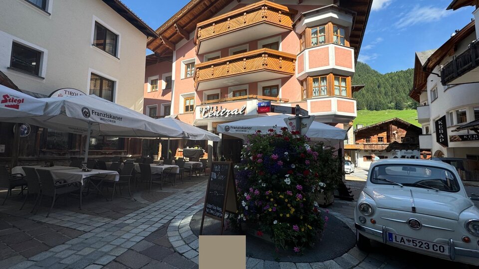 Eine malerische Stadtstraße mit charmanten Gebäuden und einem kleinen Platz. Auf dem Platz stehen Blumen und ein klassisches Auto parkt in der Nähe.
