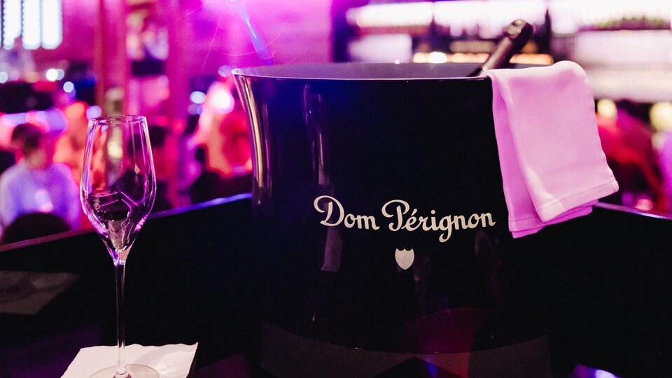 Eine Flasche Dom Pérignon in einem eleganten Eiskübel steht bereit. Im Hintergrund ist eine festliche Atmosphäre mit einem unscharfen Publikum zu sehen.