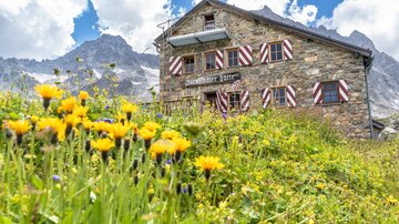 Eine malerische Berghütte aus Stein, umgeben von bunten Blumen und herrlicher Natur. Im Hintergrund sind majestätische Berge und ein blauer Himmel zu sehen.