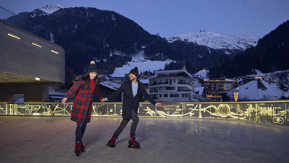 Ein Paar schlägt auf einer Eislaufbahn in den Bergen eine flotte Runde. Im Hintergrund sind schneebedeckte Gipfel und gemütliche Häuser zu sehen.