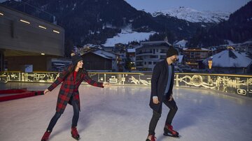 Zwei Personen skate auf einer Eislauffläche bei Abenddämmerung. Im Hintergrund sind schneebedeckte Berge und gemütliche Häuser zu sehen.