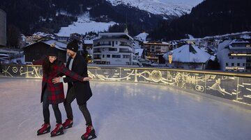 Ein Paar steht auf einer Eisbahn und genießt das Schlittschuhlaufen. Im Hintergrund sind schneebedeckte Berge und winterliche Gebäude sichtbar.