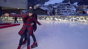Eine Eislaufbahn mit fröhlichen Menschen, die zusammen Spaß haben. Im Hintergrund sind schneebedeckte Berge und winterliche Häuser sichtbar.
