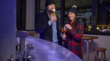 Ein Paar steht an einer Bar und genießt Getränke. Beide tragen warme Mützen und die Atmosphäre ist gemütlich.