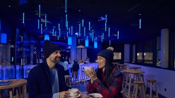 Ein Café mit zwei Personen, die in warmen Winterkleidung sitzen und sich unterhalten. Die Umgebung ist in blauem Licht beleuchtet, und sie genießen heiße Getränke.