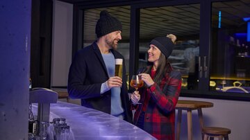 Ein Mann und eine Frau stehen an einer Bar und stoßen mit ihren Getränken an. Beide tragen Mützen und lächeln sich an.