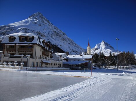 Ein malerisches Bergdorf im Schnee mit majestätischen Bergen im Hintergrund. Die Sonne scheint klar am blauen Himmel. | © TVB Paznaun -  Ischgl