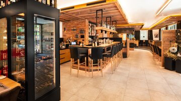 Eine moderne Bar mit Holzdetails und einer langen Theke. Hinter der Theke stehen Weinflaschen und es gibt gemütliche Sitzplätze.