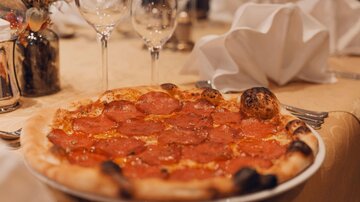 Eine lecker belegte Pizza mit Salami auf einem eleganten Tisch. Im Hintergrund stehen Gläser und Servietten bereit.