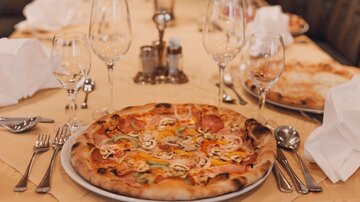 Ein Tisch mit elegantem Geschirr und Gläsern. Im Vordergrund liegt eine große Pizza mit verschiedenen Belägen.