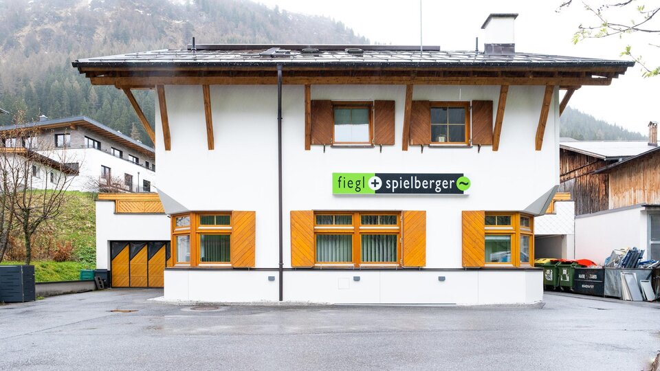 Ein modernes Gebäude mit einem weißen Außenanstrich und Holzverkleidungen. Das Logo von "fiegL spielberger" ist gut sichtbar über dem Eingang angebracht.