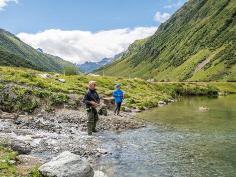 Two fishermen stand on the shore of a clear body of water and fish. The surroundings are green and inviting.