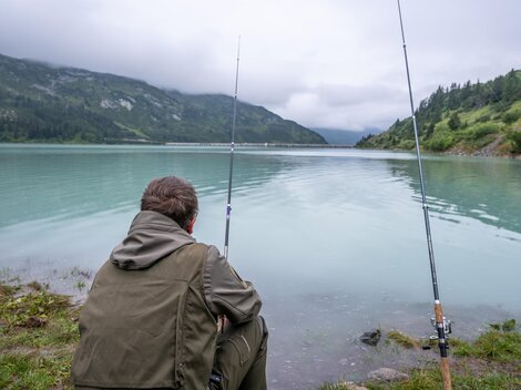 Two anglers stand by the shore of a clear mountain lake. Surrounded by green mountains and blue sky, they enjoy nature.