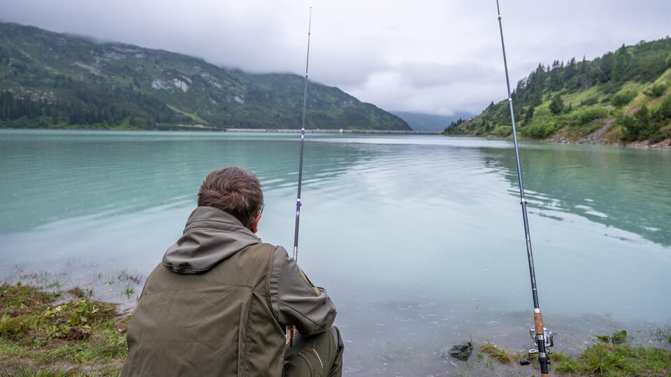 Zwei Angler stehen am Ufer eines klaren Bergsees. Umgeben von grünen Bergen und blauem Himmel genießen sie die Natur.