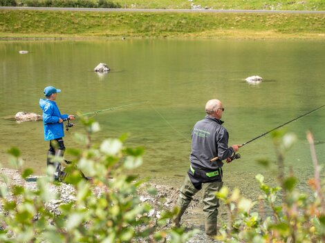 Two anglers are standing by the shore of a clear body of water. In the background, green meadows and stones can be seen.