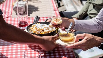 Ein Tisch mit kariertem Tischdecken, auf dem zwei Personen Essen und Getränke teilen. Es gibt eine Schüssel mit Gerichten und Gläser mit verschiedenen Getränken.