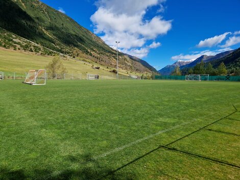 A green football field in a picturesque mountain landscape. In the background, high mountains and a clear blue sky can be seen.