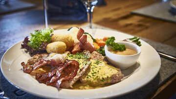 Ein Teller mit herzhaftem Essen, darunter Fleisch, Speck und Gemüse. Daneben gibt es eine leckere Sauce und Kartoffeln.