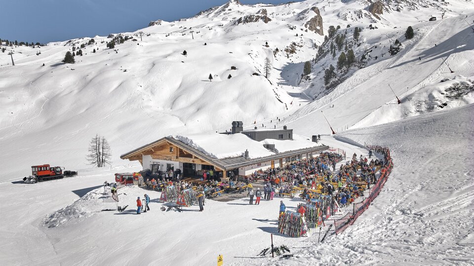 Ein belebtes Skigebiet mit einem großen Restaurant inmitten schneebedeckter Berge. Viele Menschen genießen die Sonne und die Winteraktivitäten.