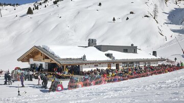 Ein Skigebiet mit einem großen, schneebedeckten Gebäude. Viele Menschen sind außen versammelt und genießen den Wintertag.