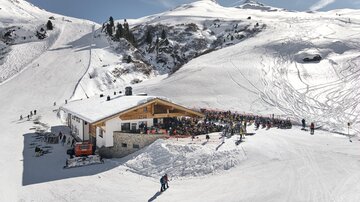 Eine Skihütte umgeben von schneebedeckten Bergen. Viele Menschen genießen die Sonne und die winterliche Atmosphäre.