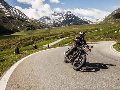 A motorcyclist rides on a winding road in the mountains. The landscape is green with snow-capped peaks in the background.