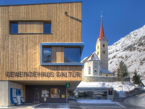 A modern community house with a wooden facade in Galtür. In the background, a church with a red bell tower and snow-covered mountains can be seen.