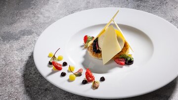 Ein kunstvoll angerichteter Teller mit einer köstlichen Speise, die frische Beeren und eine Schicht aus Käse beinhaltet. Die Dekoration umfasst bunte Fruchtpürees, die den Teller ansprechend gestalten.