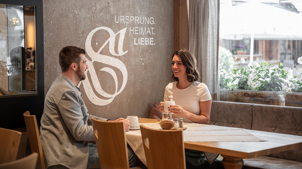 Ein gemütliches Café mit einem Paar, das miteinander spricht. An der Wand steht der Schriftzug „URSPRUNG HEIMAT LIEBE“.