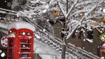Eine verschneite Szene mit einem roten Telefonzelle und einem schneebedeckten Baum. Die Umgebung ist winterlich und festlich.