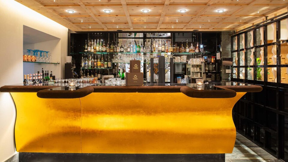 Eine elegante Bar mit einer goldenen Theke und einer Vielzahl von Spirituosen. Die Wandregale sind mit Gläsern und Getränken dekoriert.