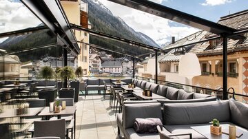 Eine elegante Dachterrasse mit bequemen Sitzgelegenheiten und Tischen. Der Ausblick umfasst wunderschöne Berge und eine charmante Stadt.