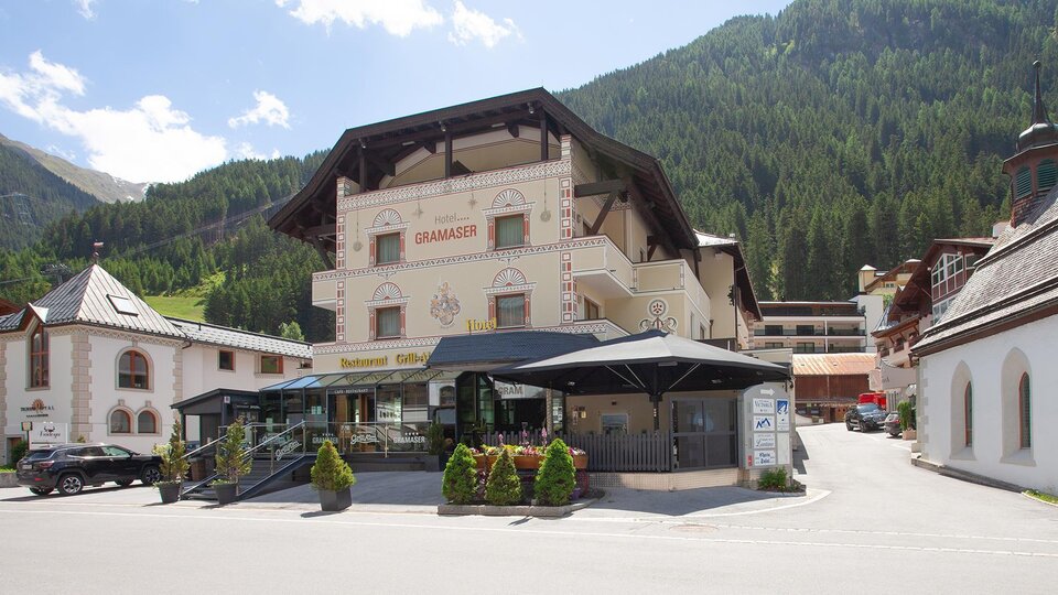 Ein malerisches Hotel im gebirgigen Umfeld mit grünen Bergen im Hintergrund. Vor dem Hotel gibt es eine gepflasterte Fläche mit Tischen und Stühlen.