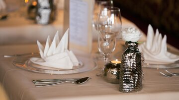 Ein elegant gedeckter Tisch mit gefalteten Servietten und Gläsern. In der Mitte steht eine stilvolle Vase mit einer Blume und einer Kerze.