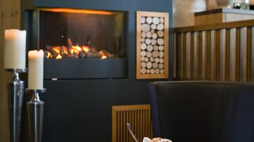 Ein gemütlicher Wohnraum mit einem modernen Kamin und Kerzenlicht. Auf dem Tisch stehen ein Getränk und ein Buch, die eine entspannte Atmosphäre schaffen.