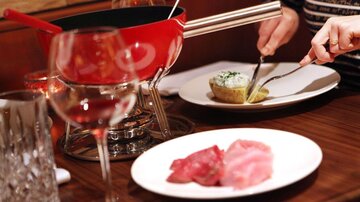 Ein Tisch mit einem roten Fondue-Topf und verschiedenen Fleischsorten auf einem weißen Teller. Im Hintergrund steht ein Glas Wein und eine Portion Kartoffel mit Kräuterquark.