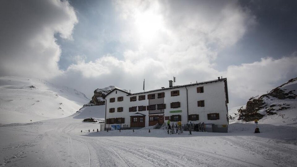 Ein weißes Gebäude im Schnee, umgeben von Bergen und Wolken. Menschen sind in der Nähe des Hauses zu sehen, das eine gemütliche Atmosphäre ausstrahlt.