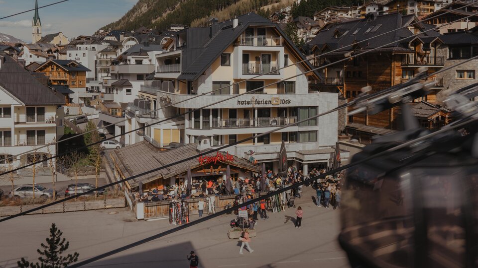Ein belebter Ort in den Bergen mit einem Hotel und vielen Menschen. Im Hintergrund sind Holzgebäude und eine Seilbahn zu sehen.