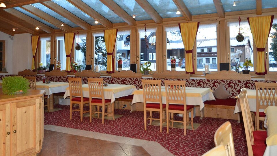 Ein gemütlicher Restaurantbereich mit Holzstühlen und Tischen. Große Fenster lassen viel Licht herein und bieten einen Blick nach draußen.