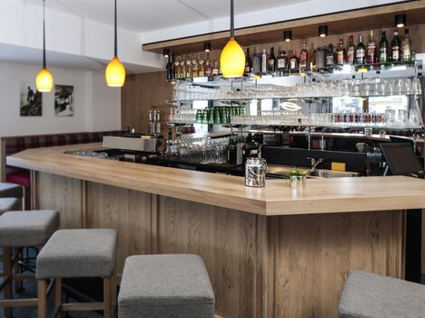 A modern bar with a curved wooden counter and stylish stools. In the background, shelves with a selection of spirits and glasses can be seen.