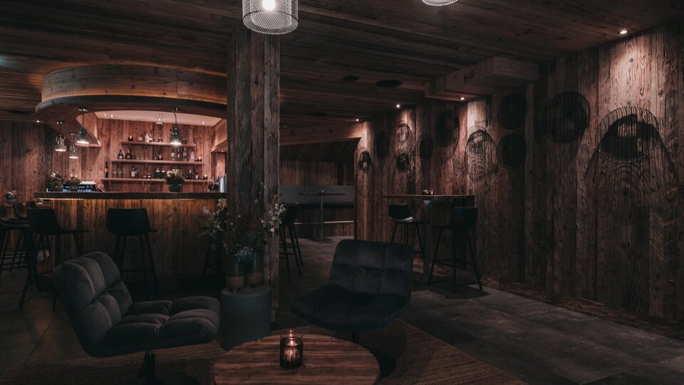 Ein gemütlicher Raum mit Holzwänden und sanfter Beleuchtung. Die Bar im Hintergrund und die modernen Möbel schaffen eine einladende Atmosphäre.
