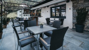 Ein stilvoller Außenbereich eines Restaurants mit Tischen und Stühlen. Im Hintergrund sind Berge und eine ruhige Umgebung zu sehen.