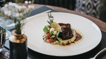 Ein elegant angerichtetes Gericht auf einem weißen Teller mit einer dunklen Sauce. Dazu frisches Gemüse und Kräuter.