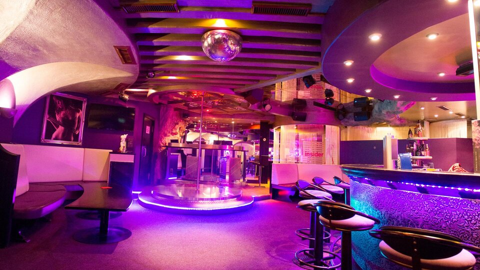 Ein stilvoller Club mit einem beleuchteten Barbereich und einer Tanzfläche in der Mitte. Die bunten Lichter schaffen eine lebhafte Atmosphäre.