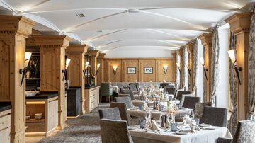 Ein elegantes Restaurant mit Holzelementen und gemütlichen Sitzplätzen. Die Tische sind festlich gedeckt und große Fenster sorgen für viel Licht.