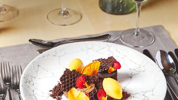 Ein elegant angerichteter Teller mit dunkler Schokolade, gelber Frucht und essbaren Blüten. Im Hintergrund sind Weingläser und eine grüne Dekoration sichtbar.