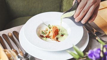 Ein elegantes Gericht wird in einen weißen Teller gegossen. Im Hintergrund sind Besteck und ein Blumengesteck zu sehen.