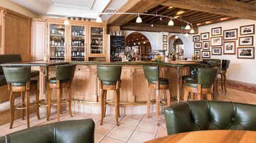 Eine stilvolle Bar mit Holzregalen und gemütlichen grünen Stühlen. An den Wänden hängen Rahmen mit vielen Fotos.